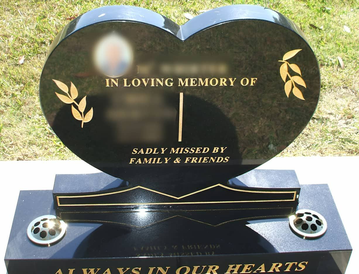 Black heart-shaped tombstone, engraved with gold text and floral accents, on a rectangular base.