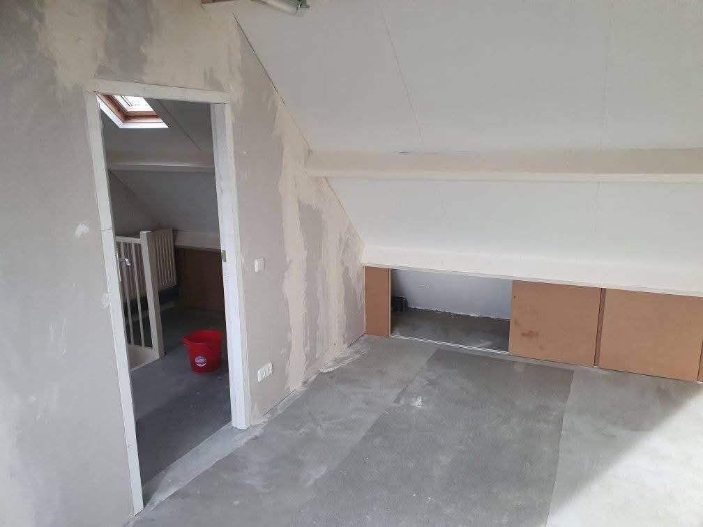 Binnenkamer met schuin plafond, deuropening, onafgewerkte muren en kartonnen opbergdozen.