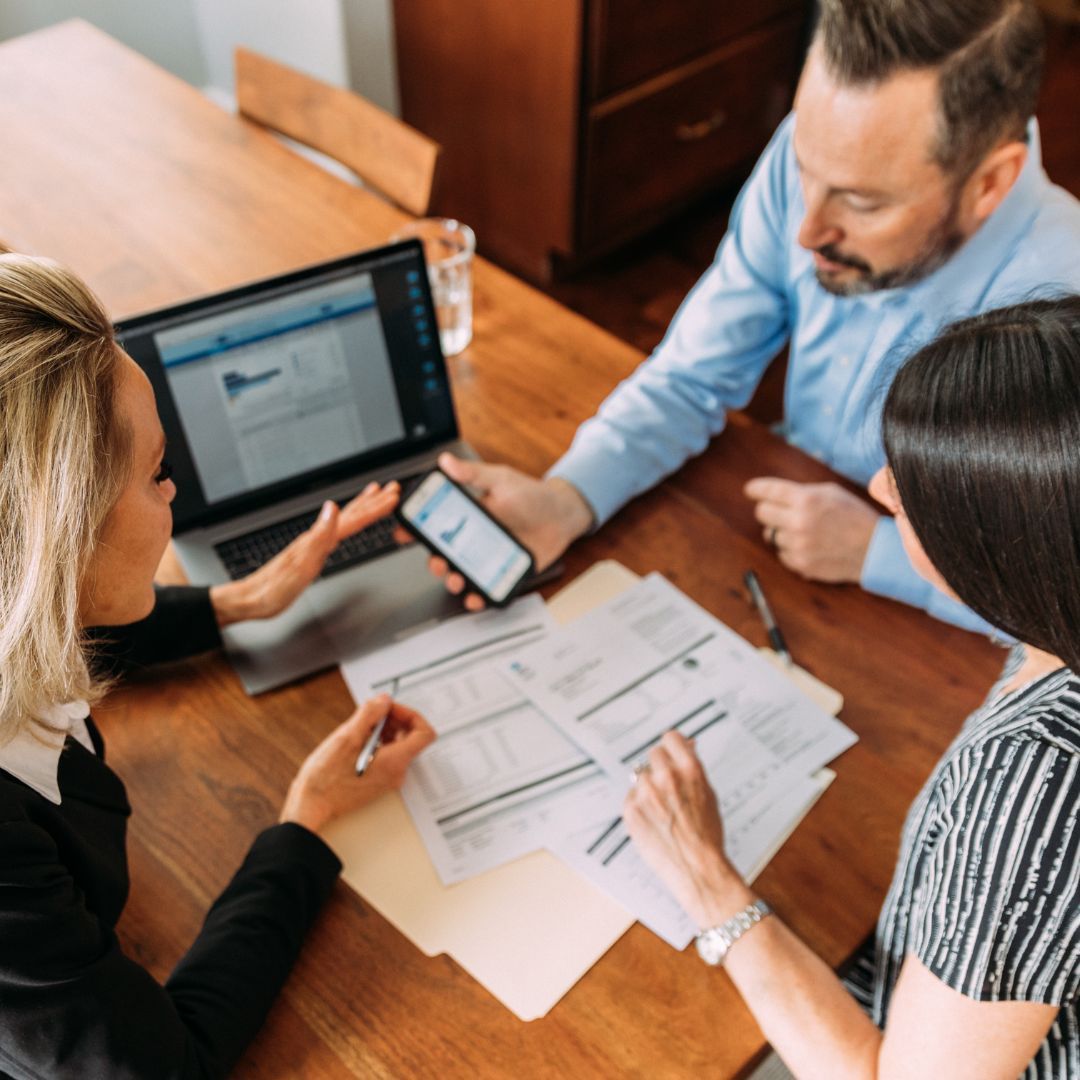 Three people reviewing documents and a laptop at a wooden table. One person holds a phone, and another points.