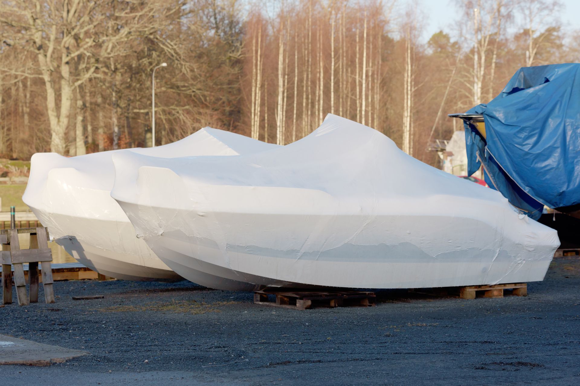 White boat hull under a cover on a trailer in an outdoor lot, with trees in the background