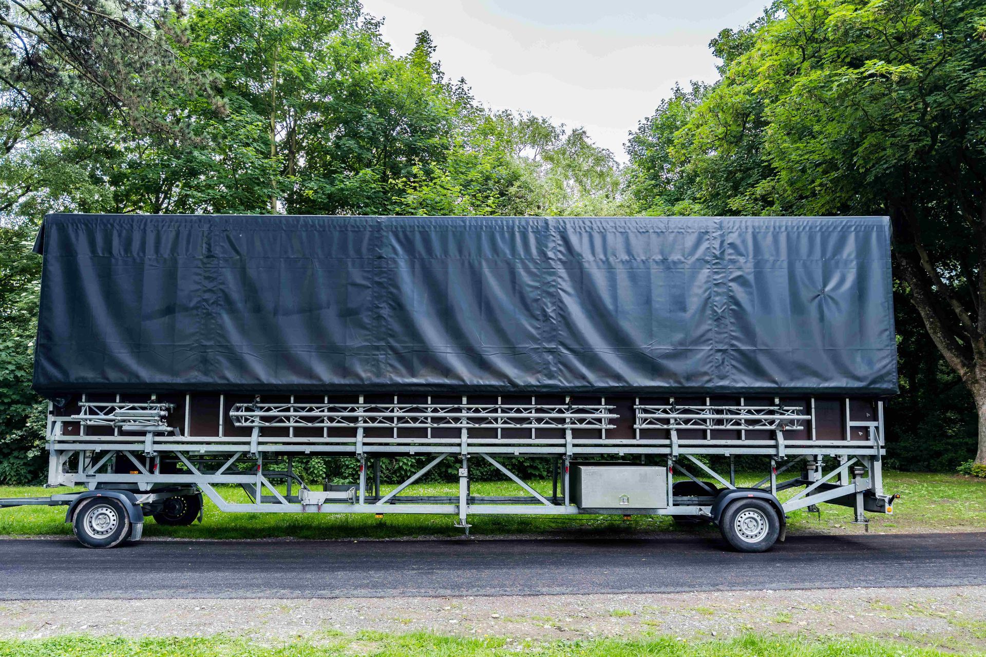 Large black tarp-covered trailer parked on a roadside with trees in the background