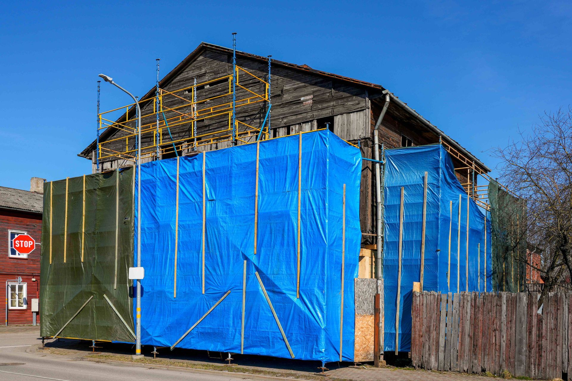 Building under construction wrapped in blue and green tarps beside a wooden fence