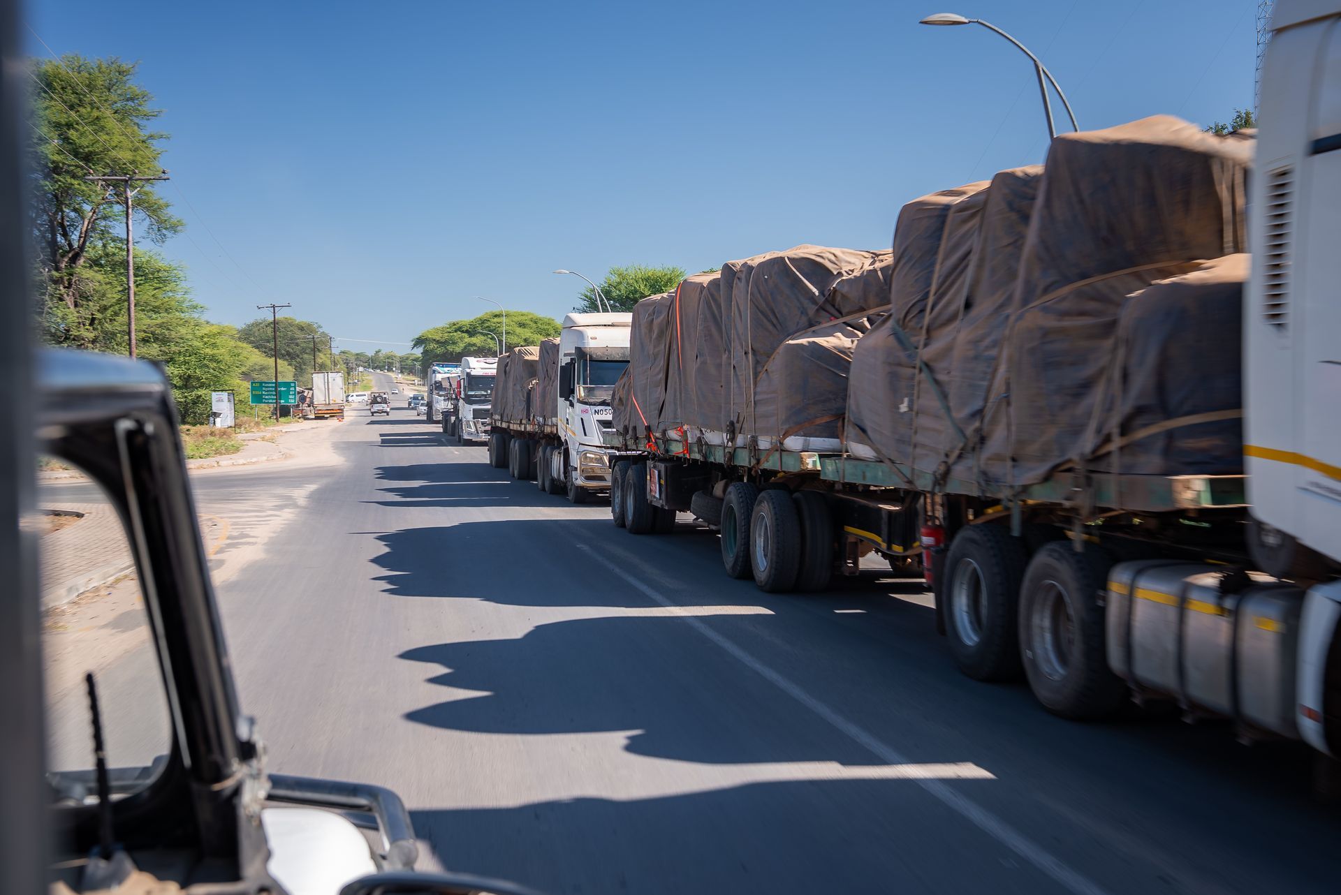 Long line of flatbed trucks with covered cargo parked along a sunny street