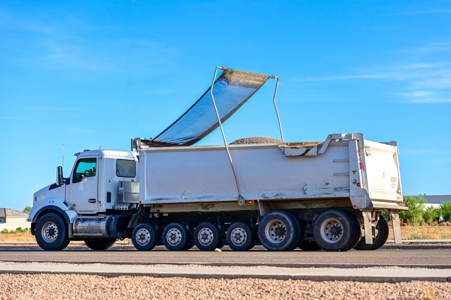 White dump truck on a road, unloading a raised load bed under a clear blue sky