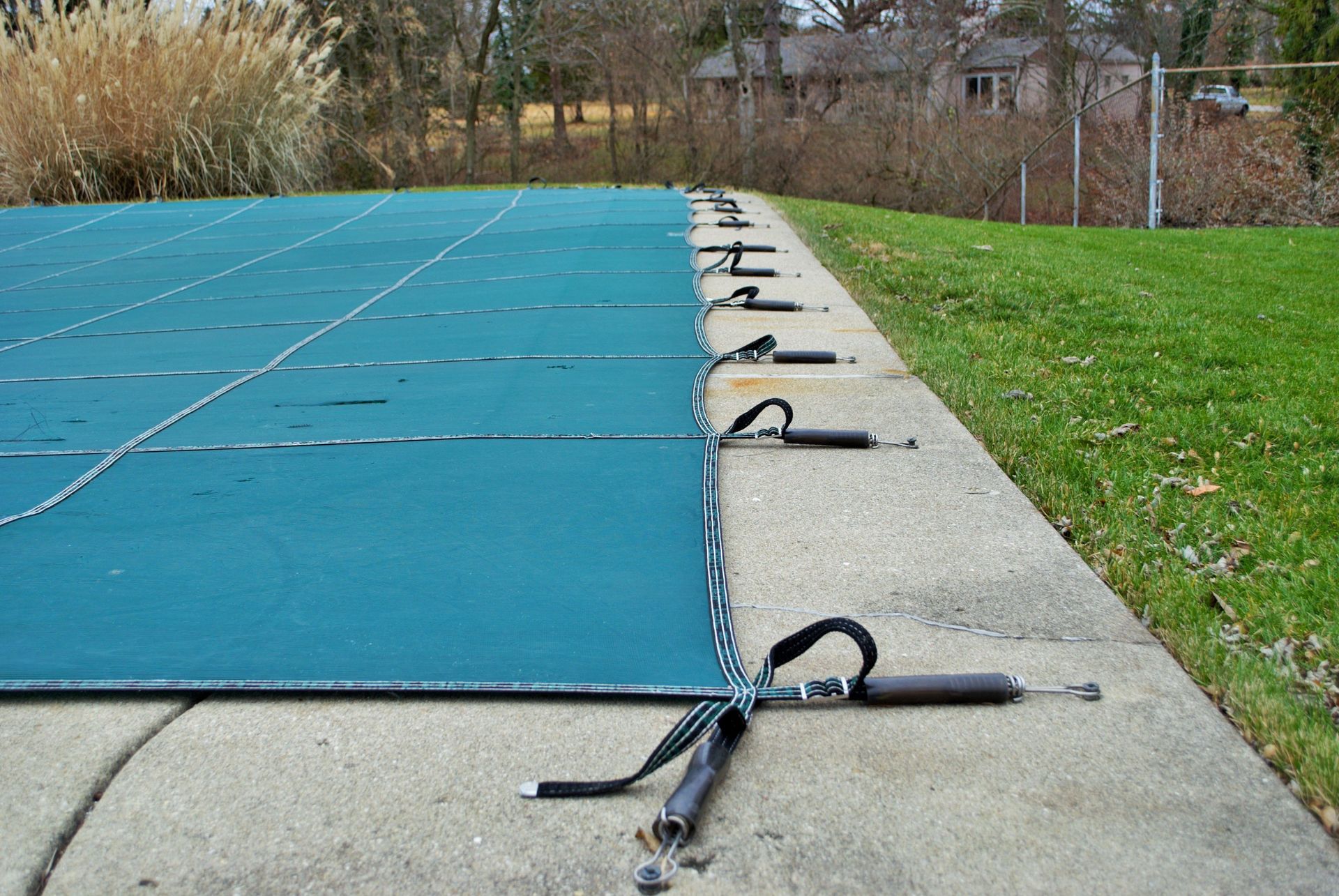 Blue covered sports court edge with straps securing a tarp beside green grass and trees