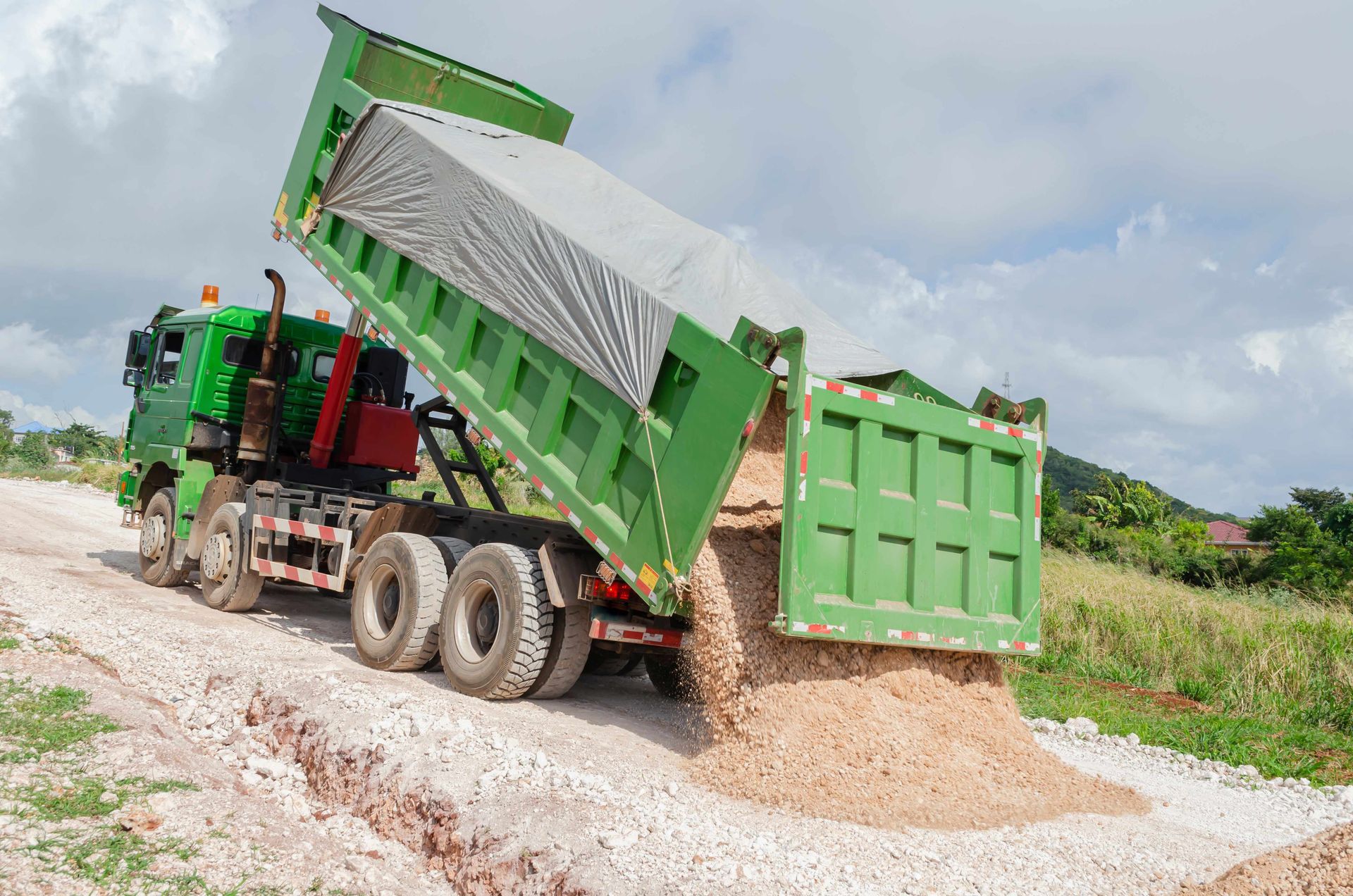 Green dump truck unloading gravel on a rural dirt road under cloudy sky