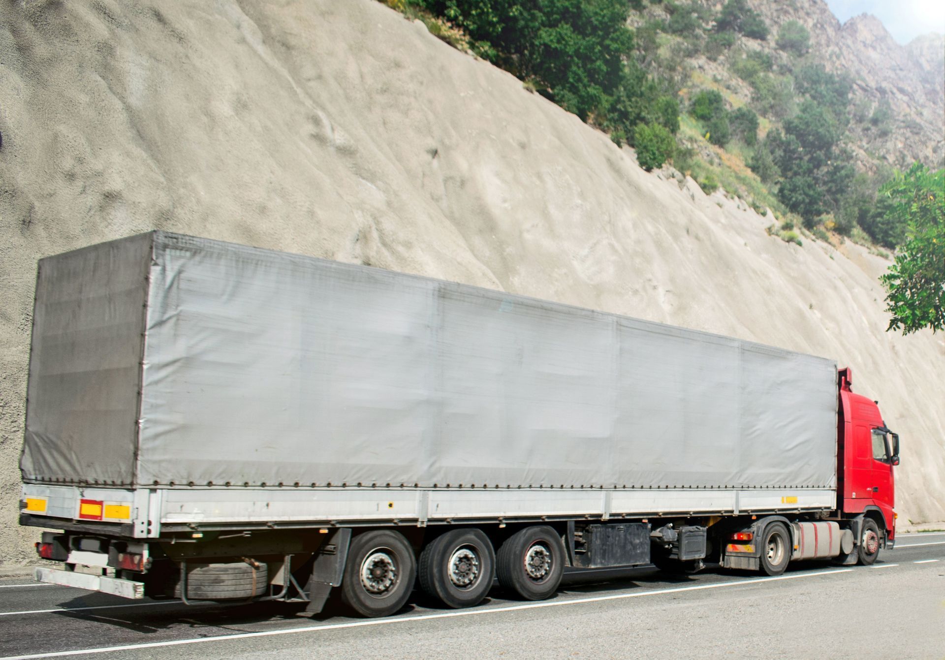 Red cargo truck with gray covered trailer parked beside a rocky roadside slope.