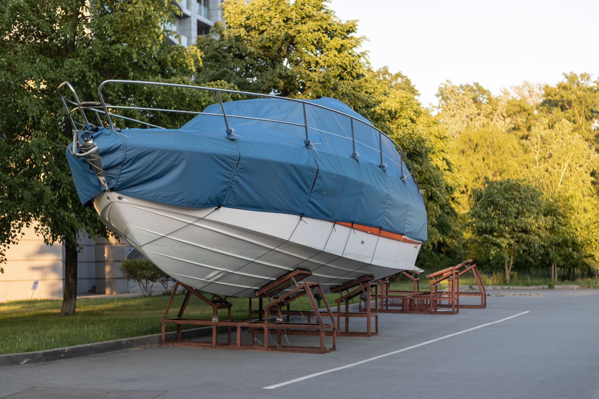 Blue sailboat on a trailer in a parking lot near trees and buildings