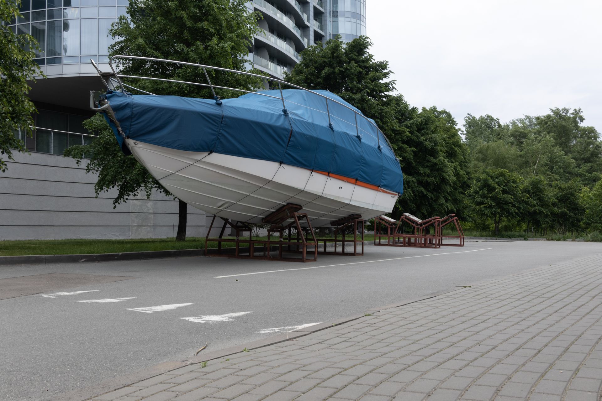 Blue-and-white sailboat sculpture on supports beside a modern building and trees