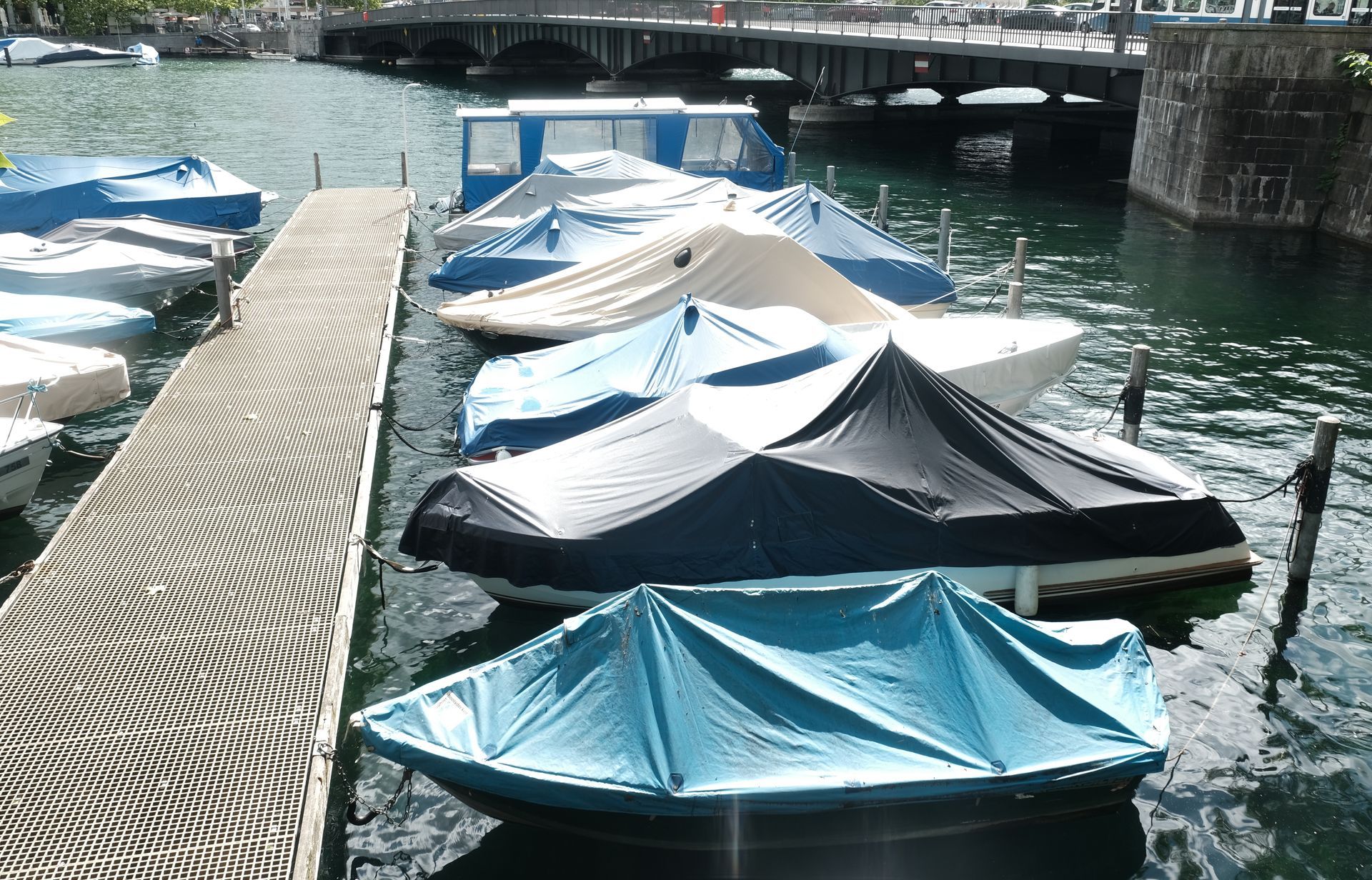 Covered boats docked along a waterfront walkway under a bridge, with blue and black protective covers.
