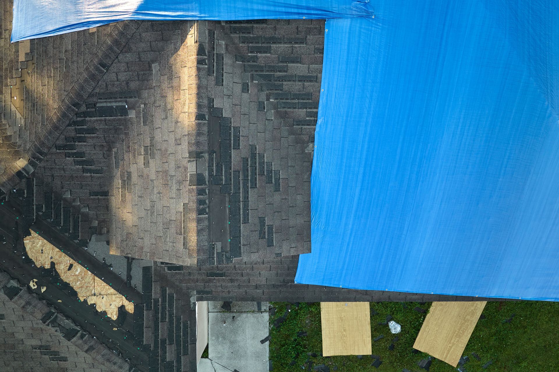 A rooftop with dark shingles beside a bright blue tarp and two tan boards on green grass.