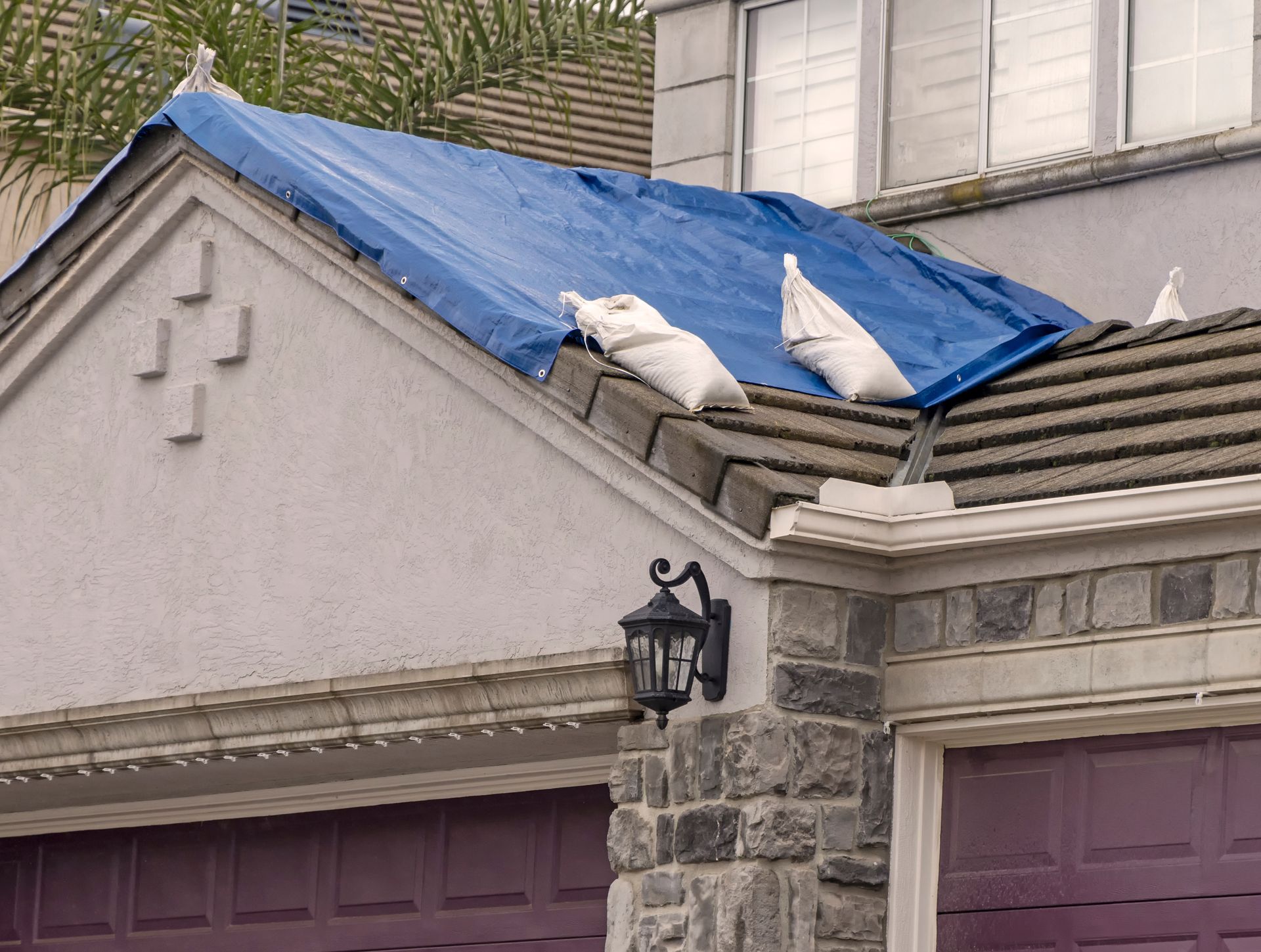 Blue tarp covering a damaged roof with sandbags on top above two garage doors Blue tarp covering a damaged roof with sandbags on top above two garage doors