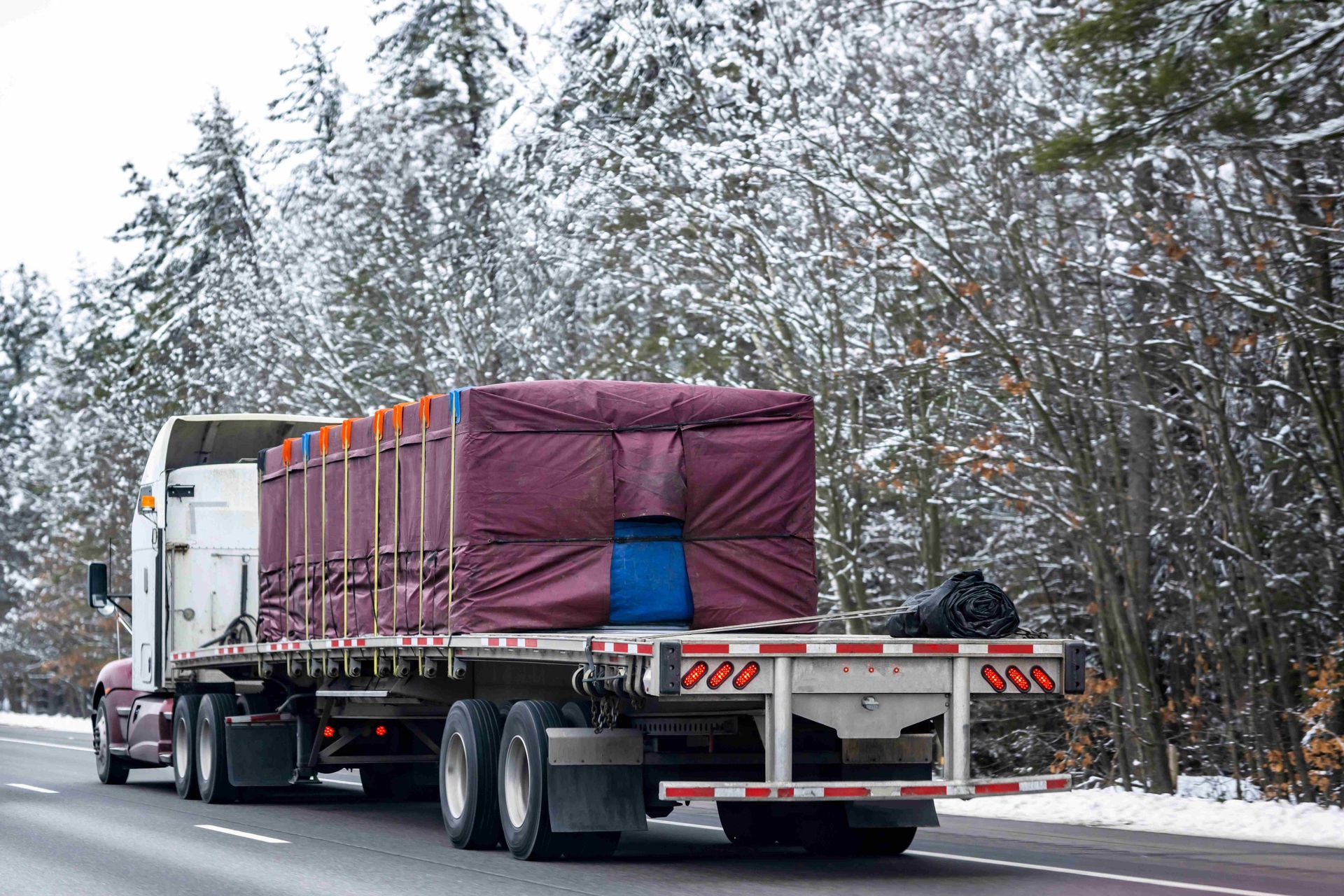 Flatbed semi truck hauling a purple-covered load on a snowy road with forest trees beside it