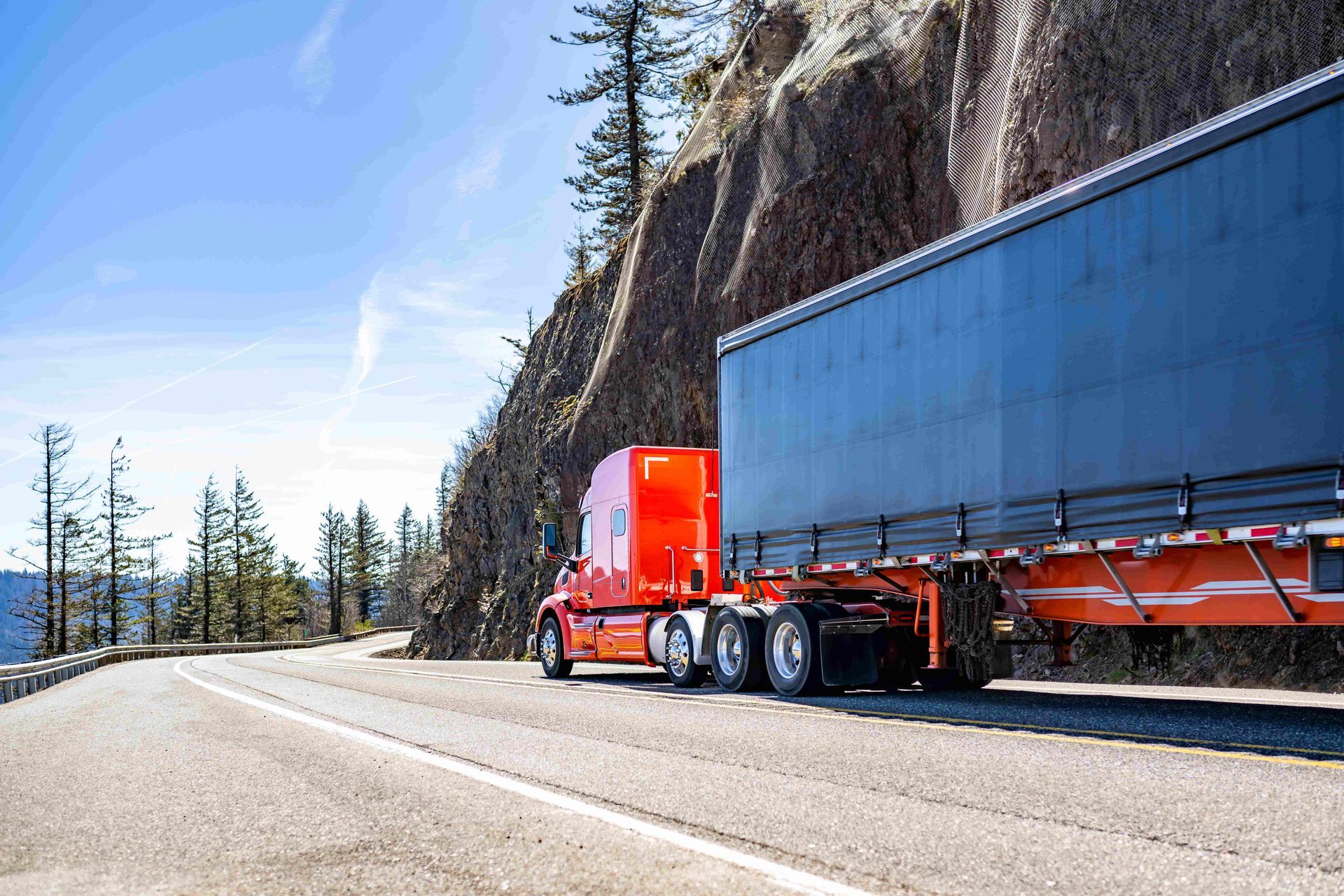 Red semi truck hauling a black trailer on a mountain road beside a rocky cliff