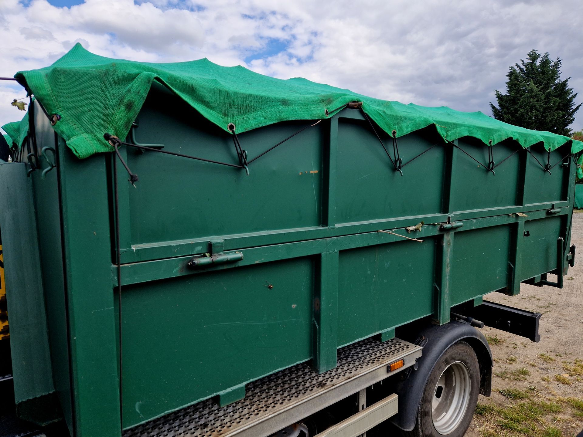 Green utility trailer with a tarp cover parked outdoors on gravel under a cloudy sky