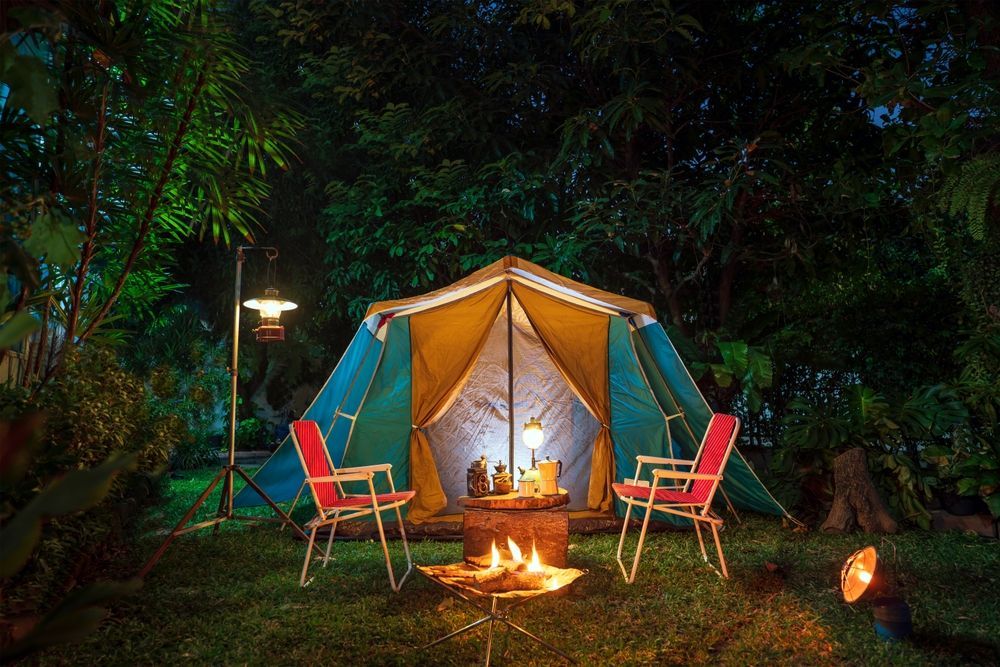A teal and yellow tent at night, set up in a wooded area with two folding chairs and a campfire glowing in the foreground.
