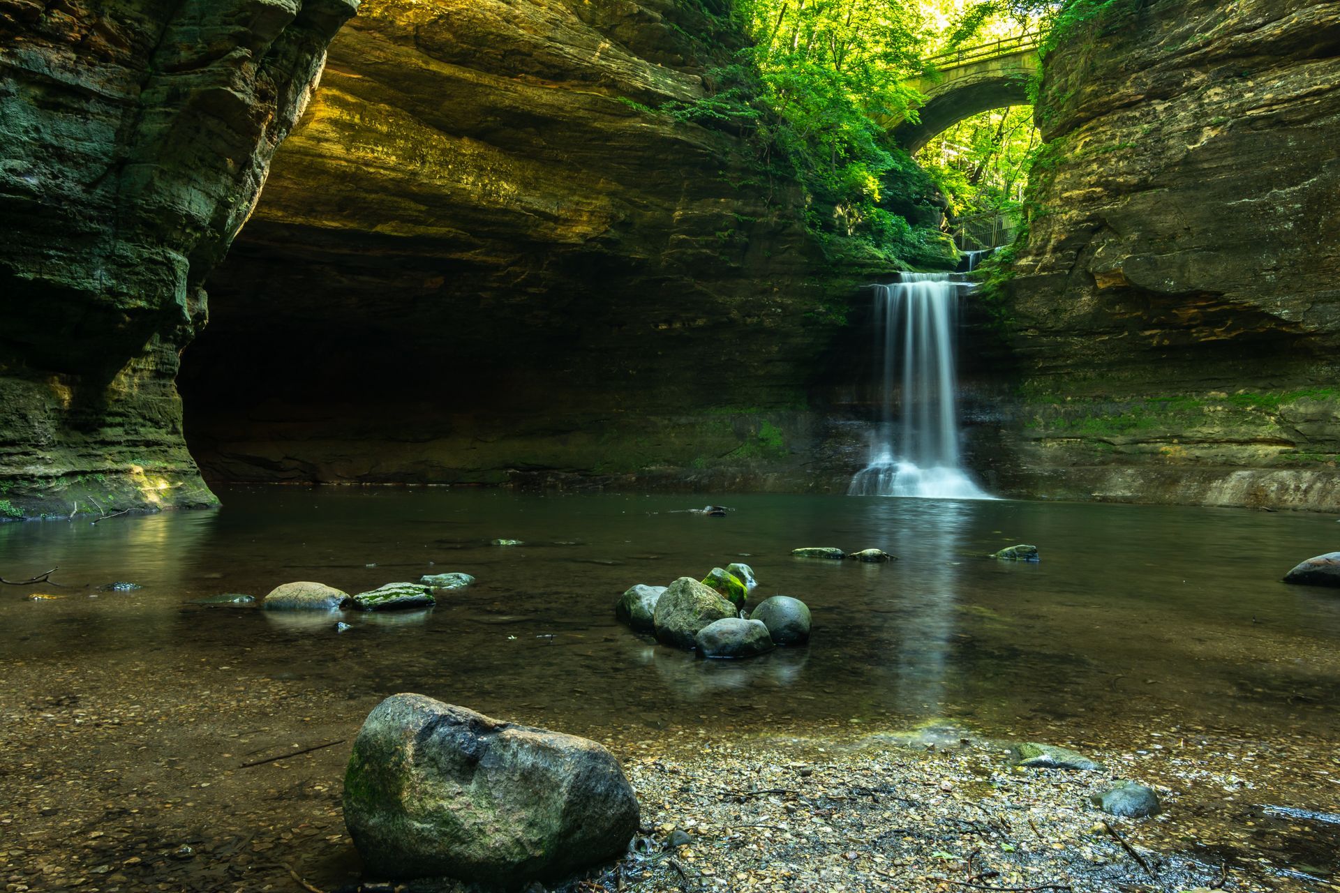 Waterfall in a rocky cave pool with mossy green cliffs and sunlight filtering through trees