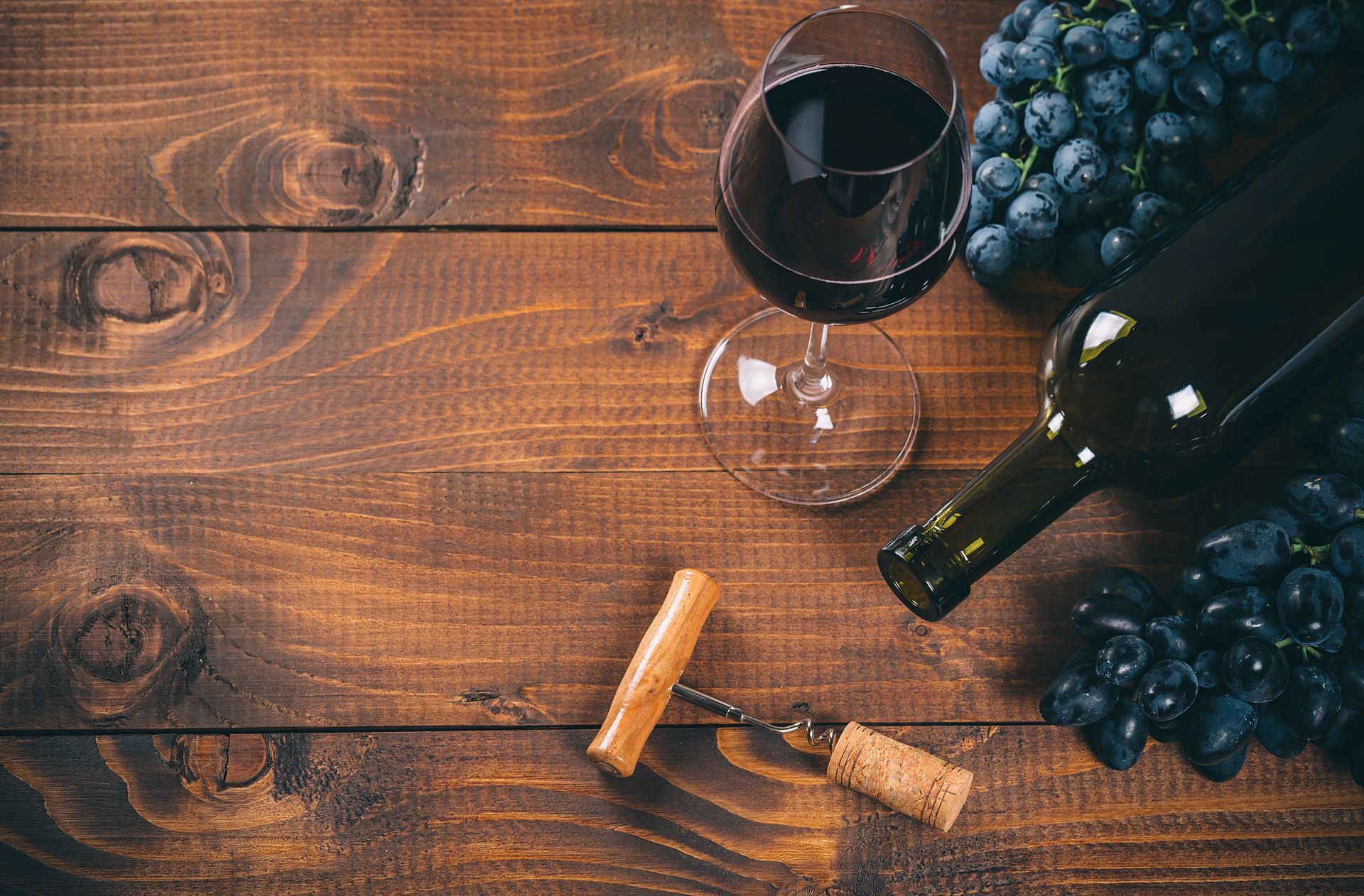 Wine bottle, glass of red wine, corkscrew, cork, and grapes on a rustic wooden table.
