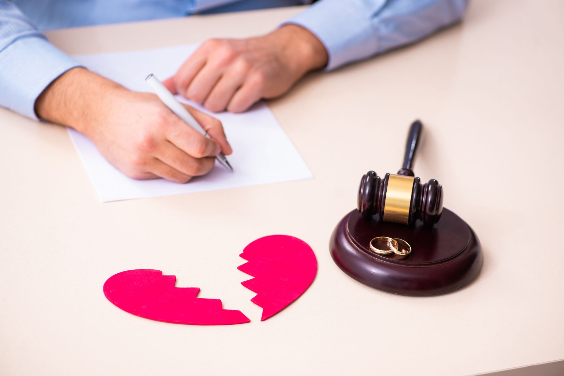 A person writing on paper, broken heart, gavel, wedding rings; divorce proceedings on a desk.