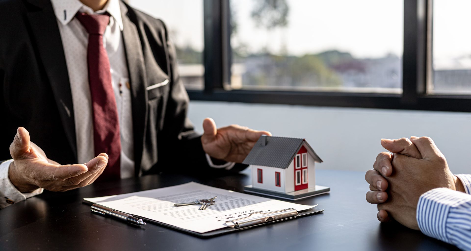 A person in a suit sits at a desk with a small house model and paperwork, discussing real estate with another person.