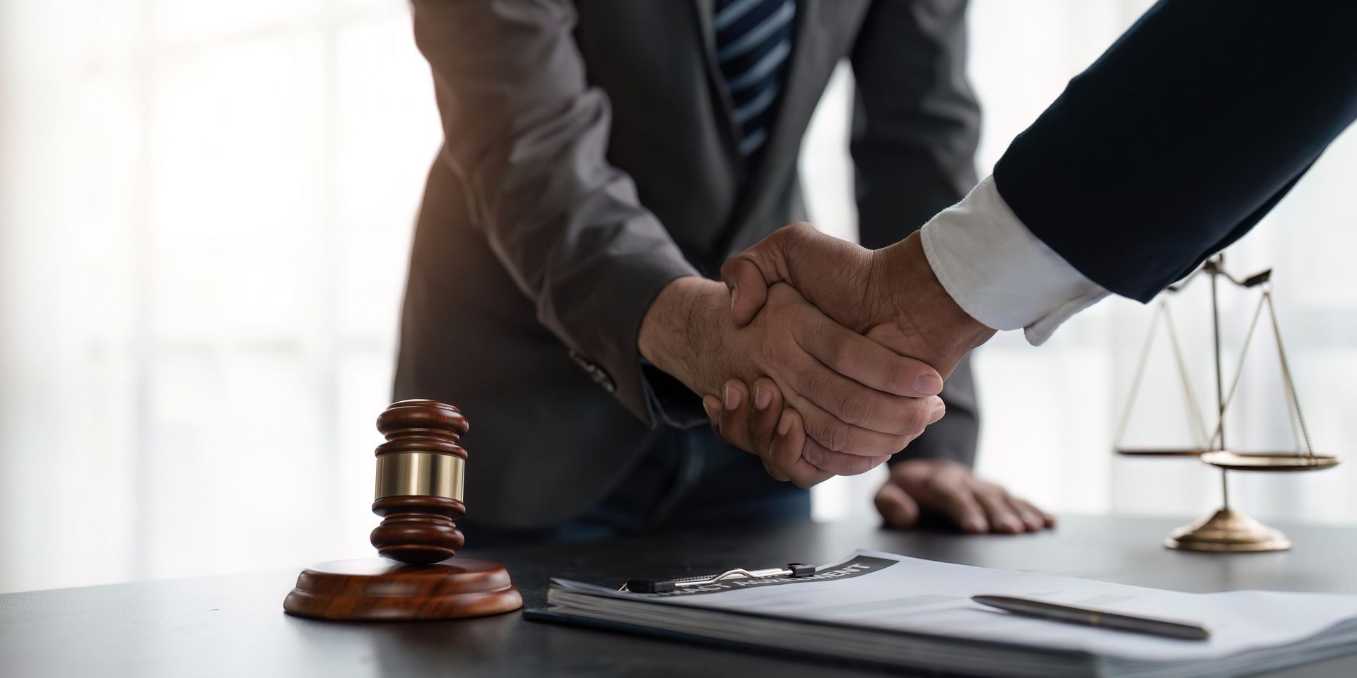 Two people shaking hands over a desk with a gavel, scales, and documents, possibly a legal setting.