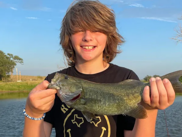 A smiling person holds a freshly caught bass in front of a lake at sunset.