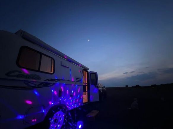 A Casita camper at twilight, illuminated by vibrant blue and purple party lights reflecting off its side.