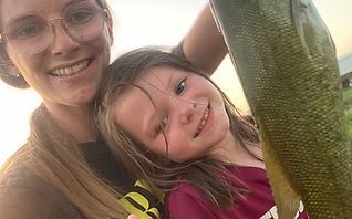 Teen holding a freshly caught fish by a pond, wearing a gray T-shirt under cloudy skies