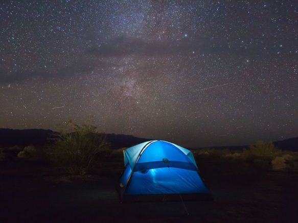 A glowing blue camping tent set against a vast night sky filled with the Milky Way galaxy.