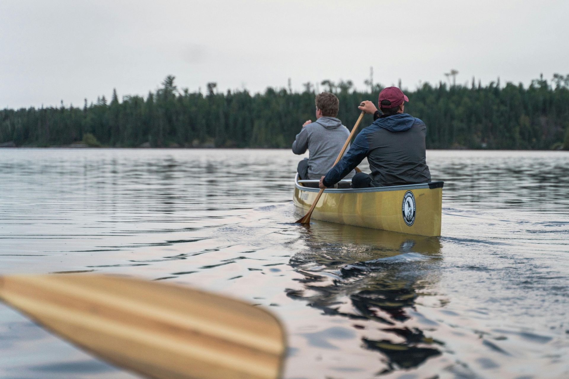 Two people canoeing on a calm lake, with a forested shoreline in the distance and a paddle in the foreground