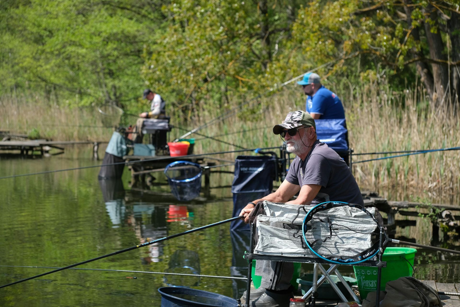 People fishing from small boats on a calm lake, with green trees along the shore