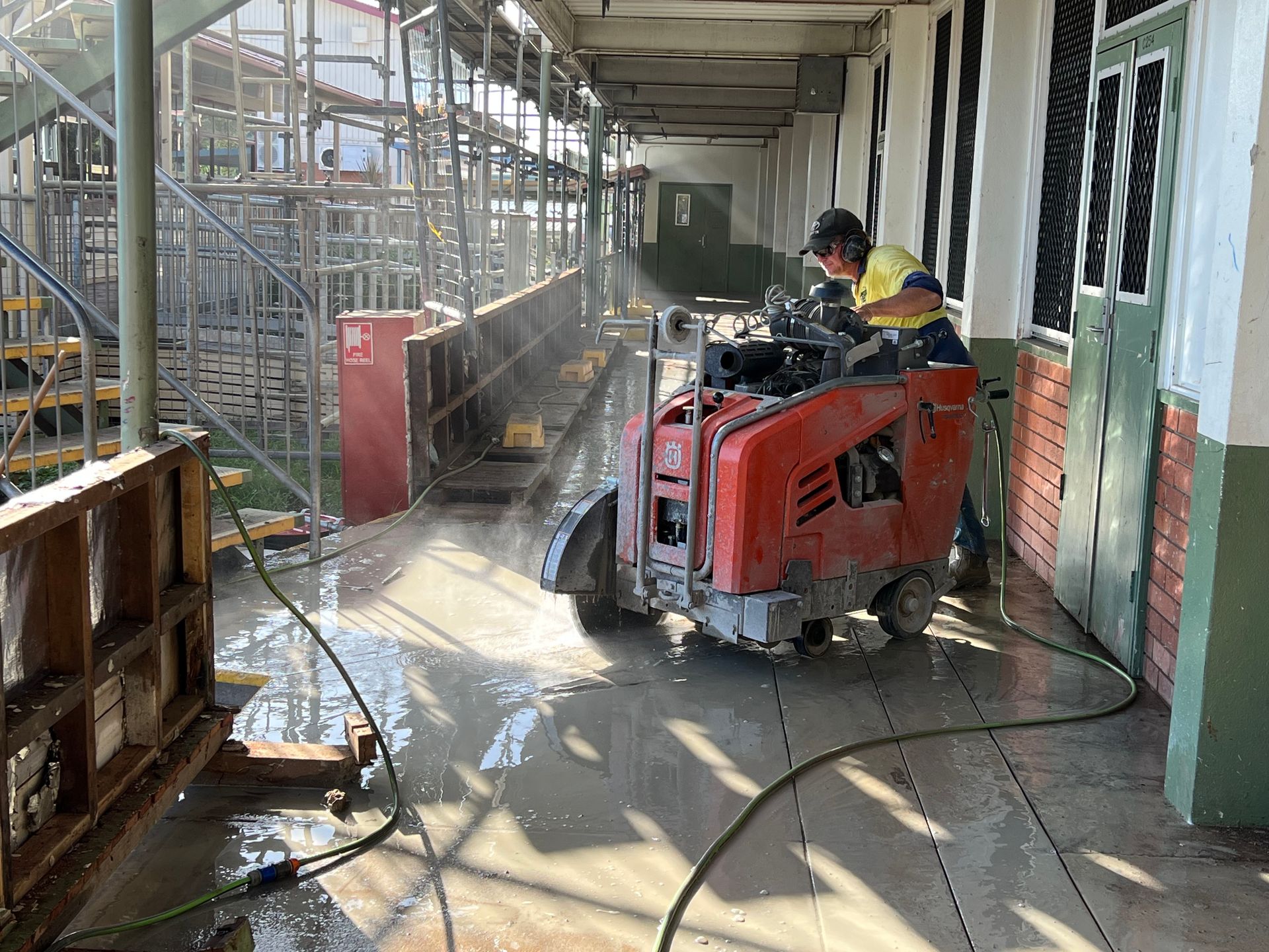 Orange and white traffic cone on sidewalk; worker operating concrete saw on street.