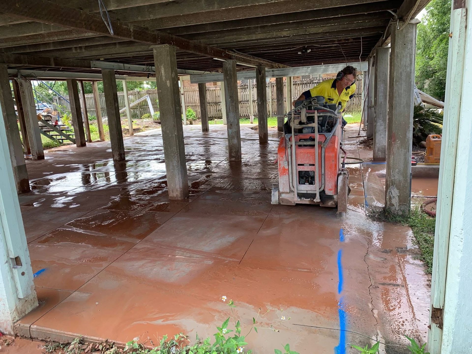 Person pressure-washing a wet concrete floor under a raised building, with water pooling and a blue hose visible — Ablecut Concrete Cutting & Coring In East Mackay, QLD