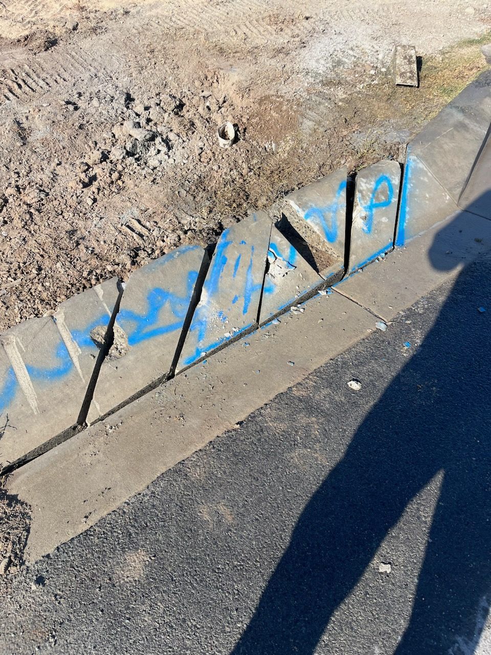 Blue graffiti on a concrete curb beside a dirt lot, with a long shadow on the pavement. — Ablecut Concrete Cutting & Coring In East Mackay, QLD