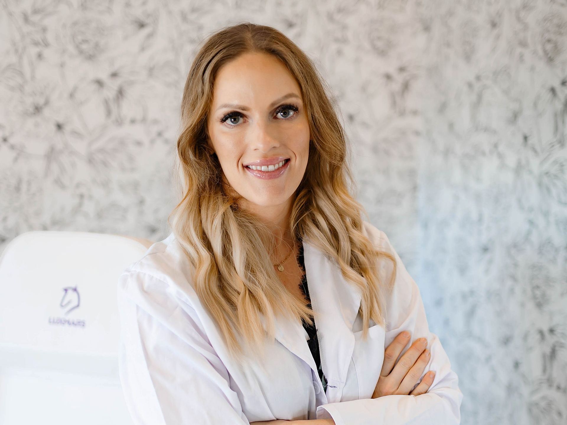 Woman in white coat smiles, arms crossed, in a light-colored medical office.