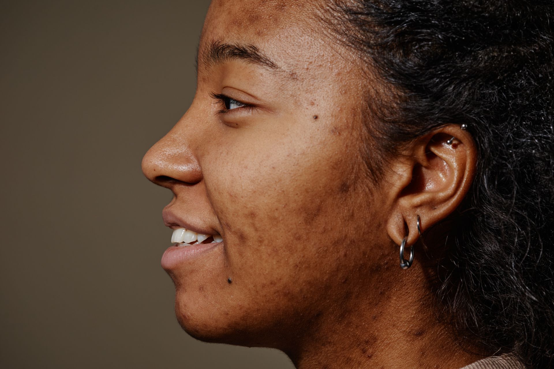 Person with dark skin, smiling, turned to the side, visible cheek and nose, wearing earrings.