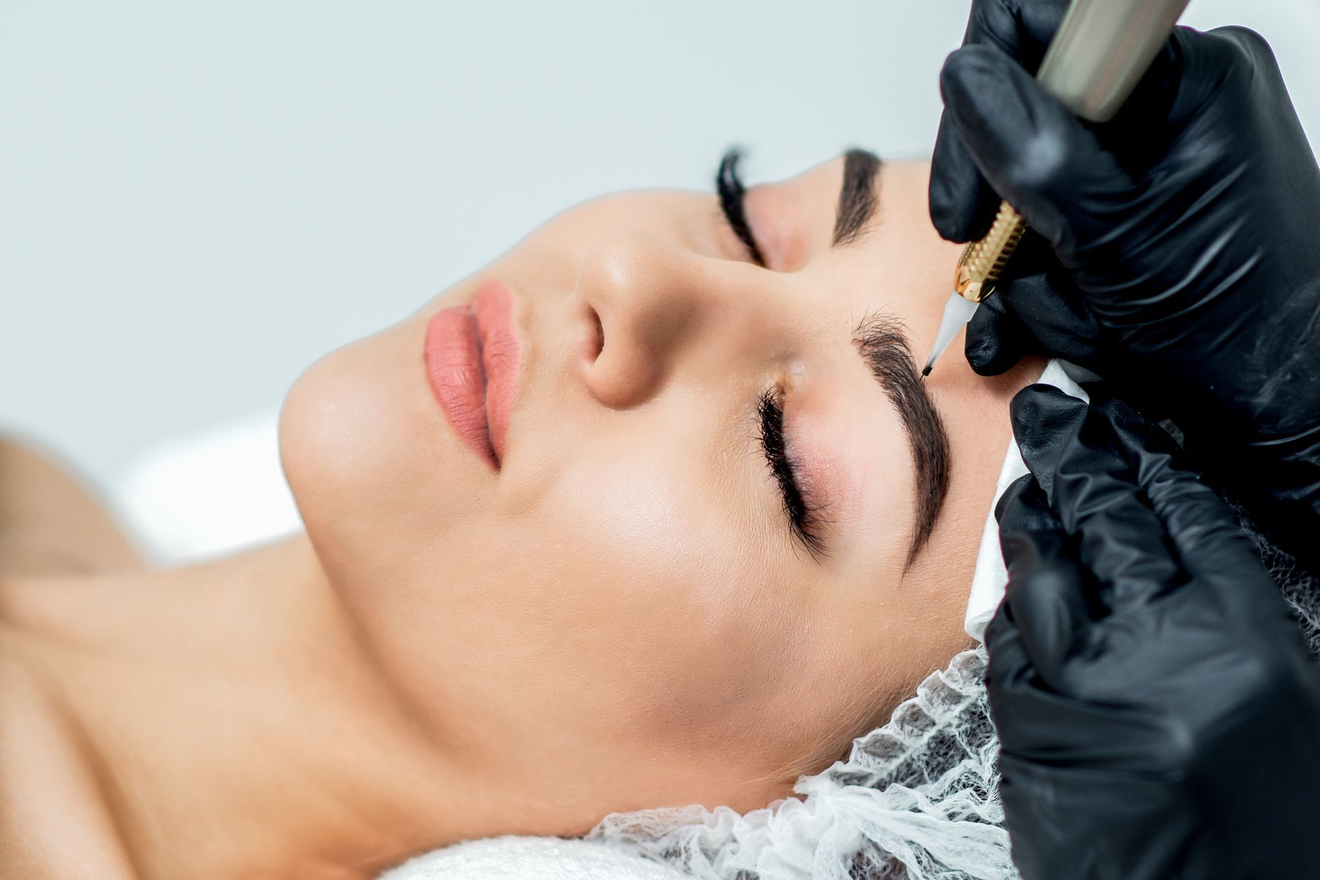 Woman receiving eyebrow microblading, eyes closed, black-gloved hands holding a tool.