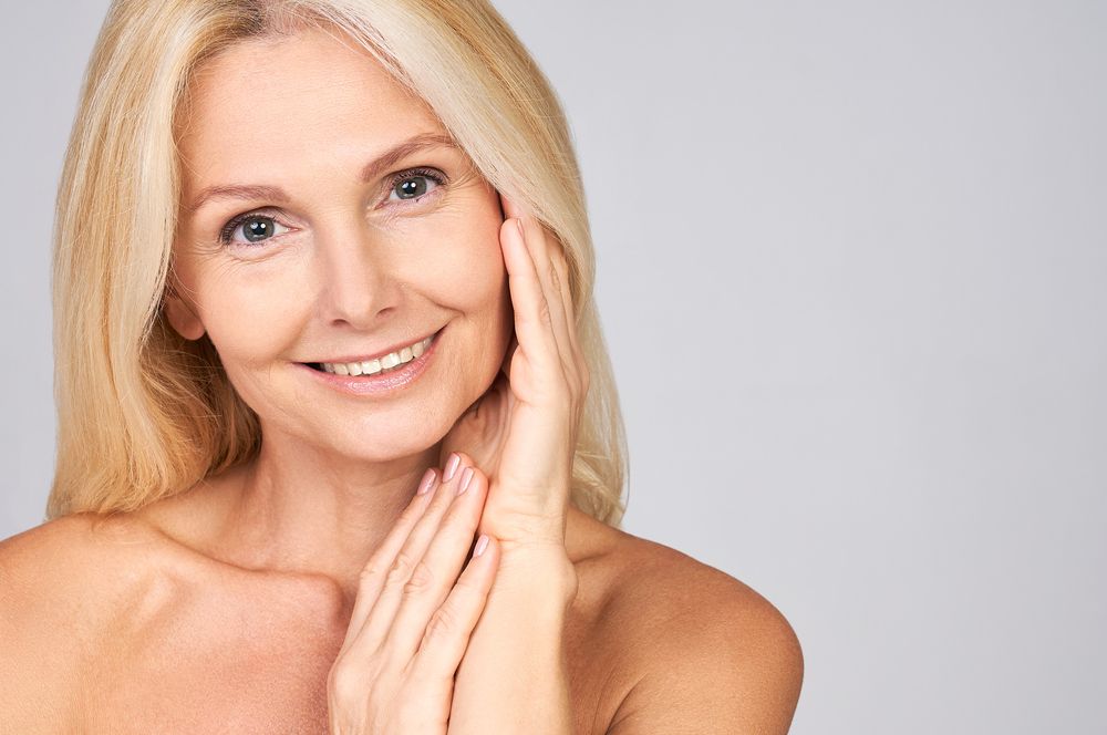 Smiling blonde woman touching her face against a light gray background