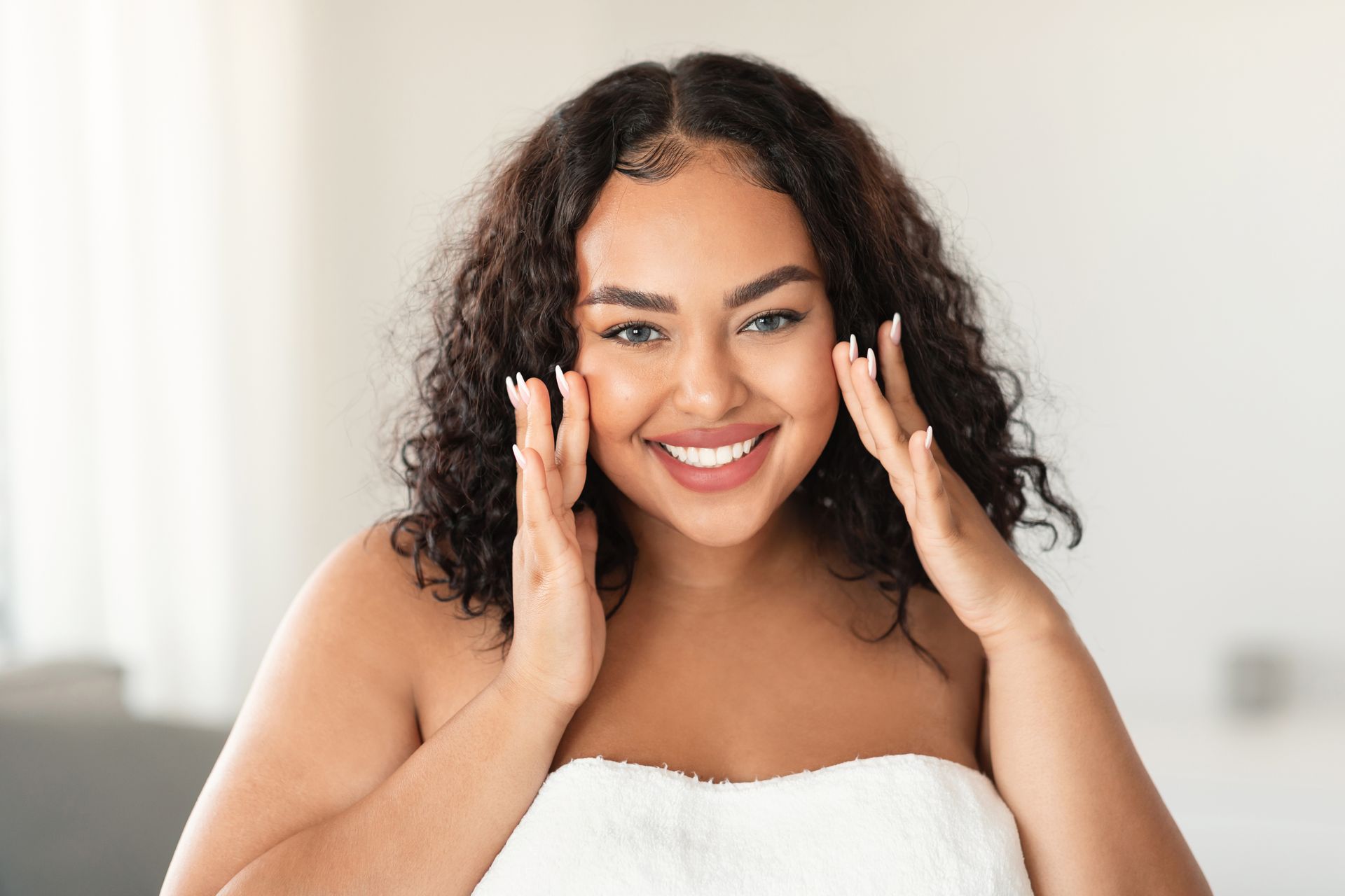 Woman with curly hair smiles, touching her cheeks with both hands, wrapped in a white towel.