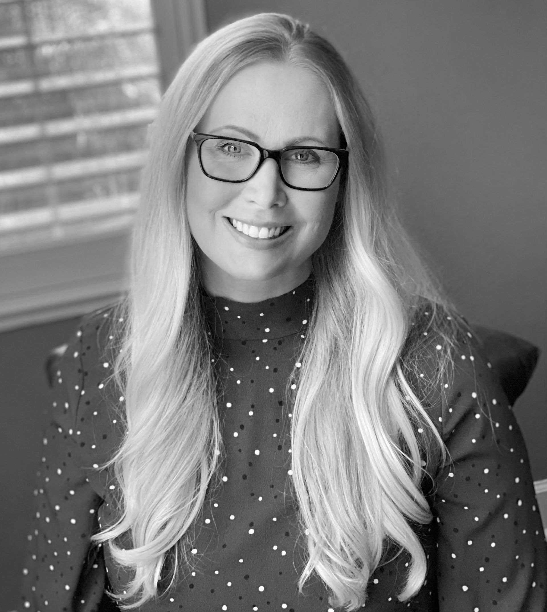 Woman with long blonde hair and glasses smiles at the camera, wearing a polka dot top. Indoor setting with blinds.
