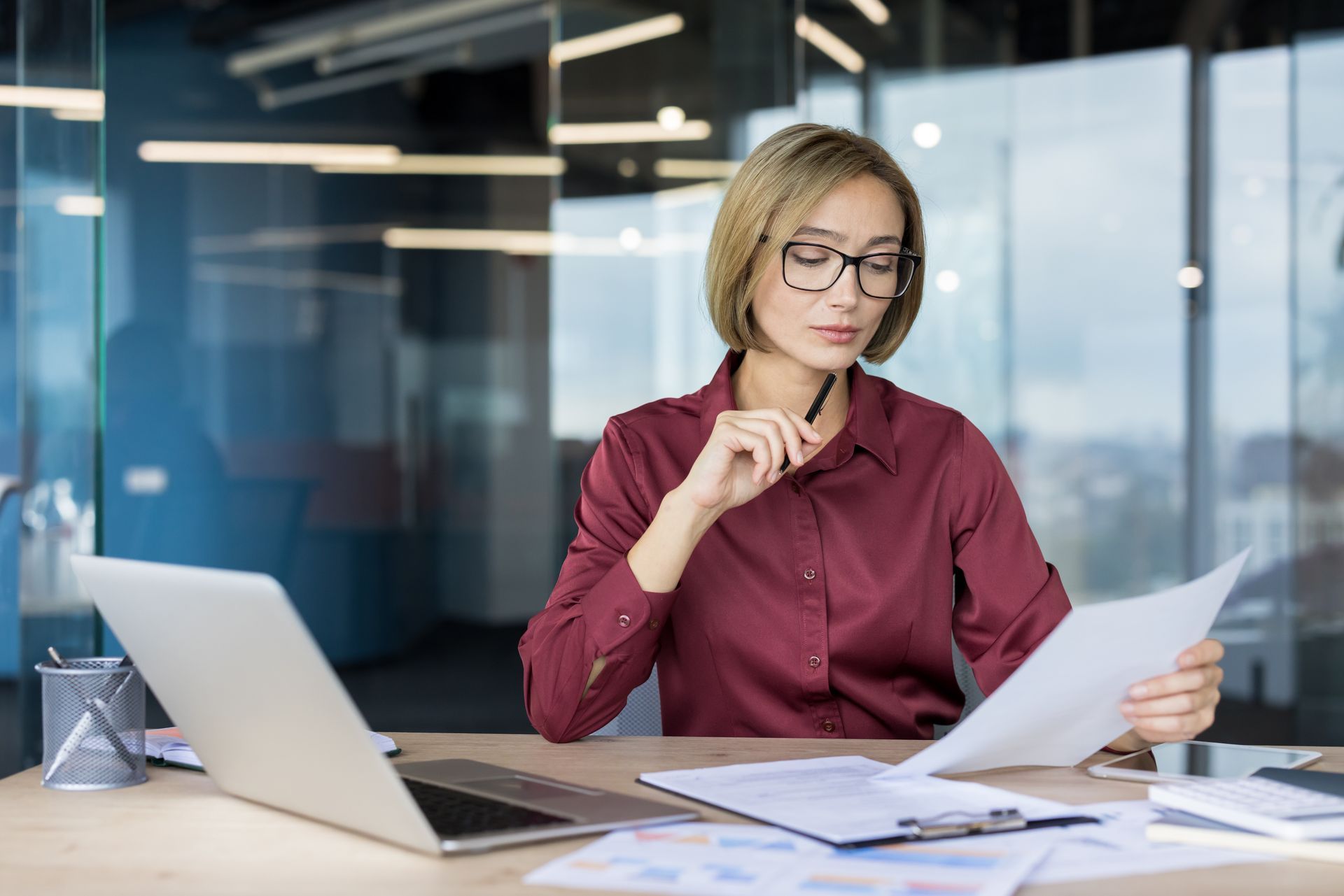 Woman in glasses reviews documents at a desk beside a laptop in a modern office.