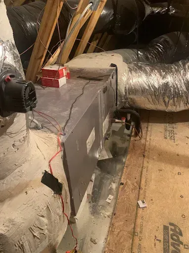 HVAC system in an attic. Metal unit with ductwork, electrical wires, and insulation. Wooden beams and flooring.