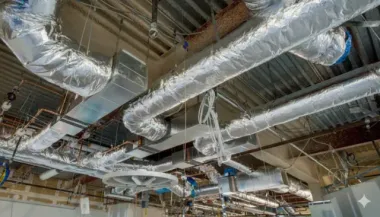 HVAC ductwork and pipes installed on a high, unfinished ceiling of a building.