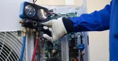 HVAC technician in blue sleeve, connecting gauges to an air conditioning unit.