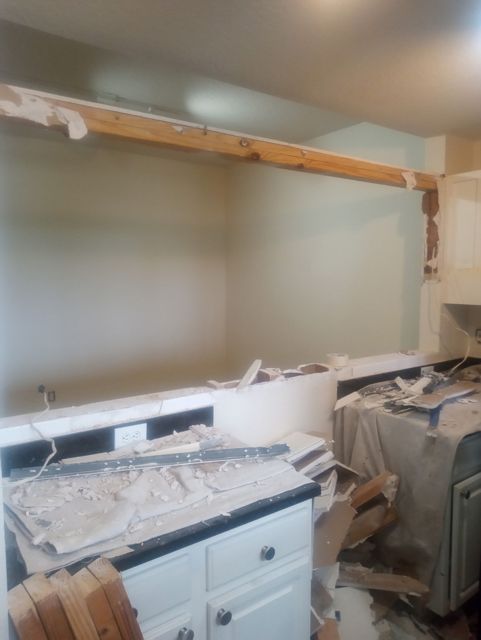 Kitchen undergoing demolition; debris on countertops and floor. Exposed wooden beam and damaged walls.