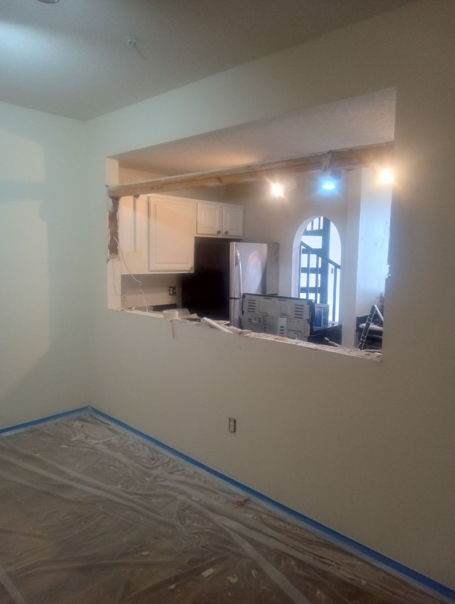Wall with a rectangular opening revealing a kitchen with white cabinets and a refrigerator.