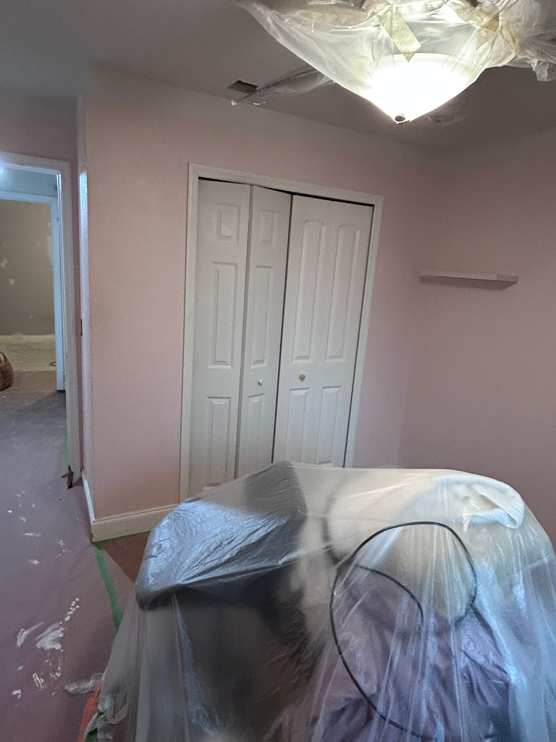 Room with pink walls, white bi-fold closet doors, and covered furniture. Ceiling light covered in plastic.