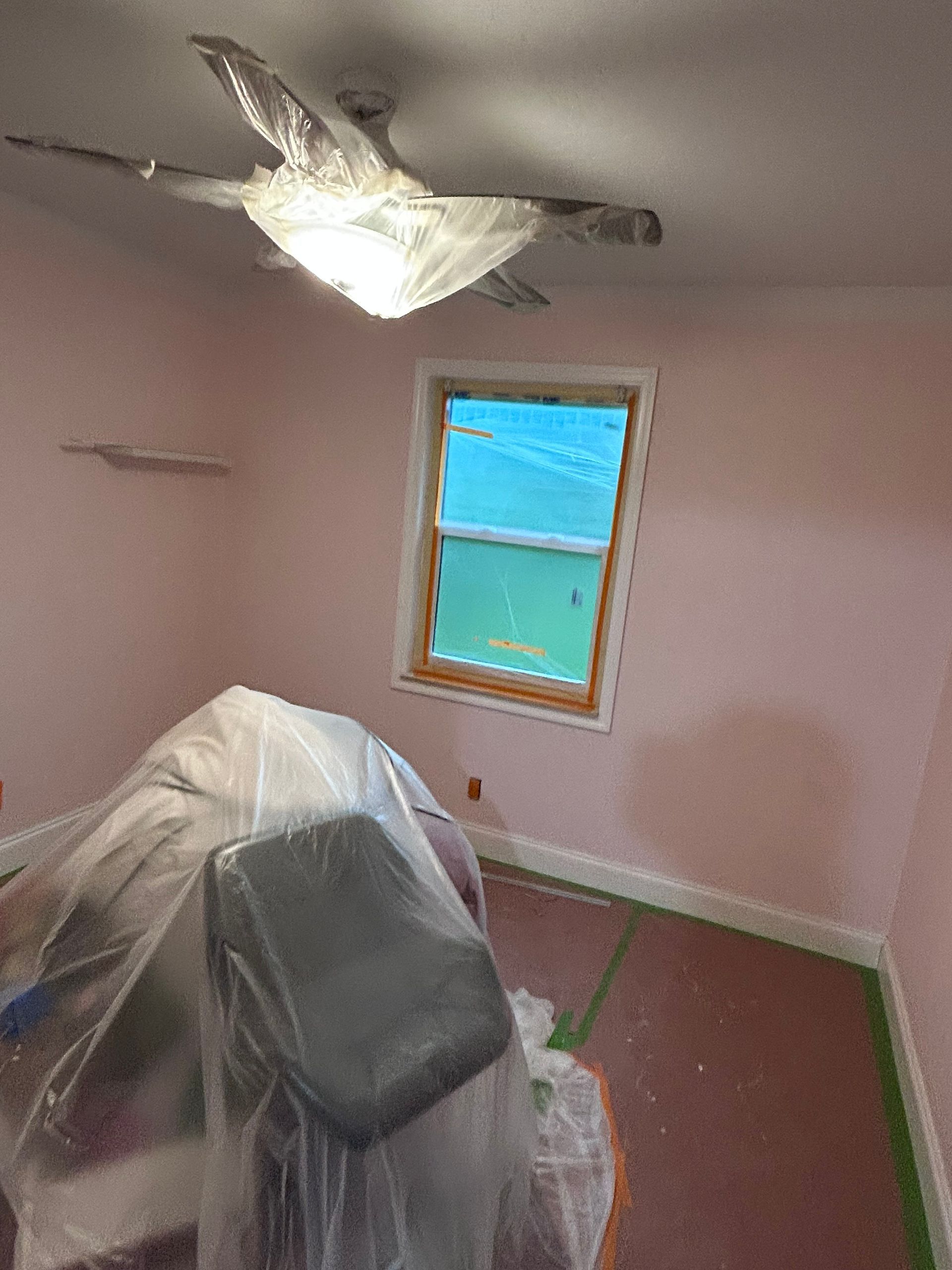 Room with pink walls, window, and ceiling fan covered in plastic. Furniture is also covered.