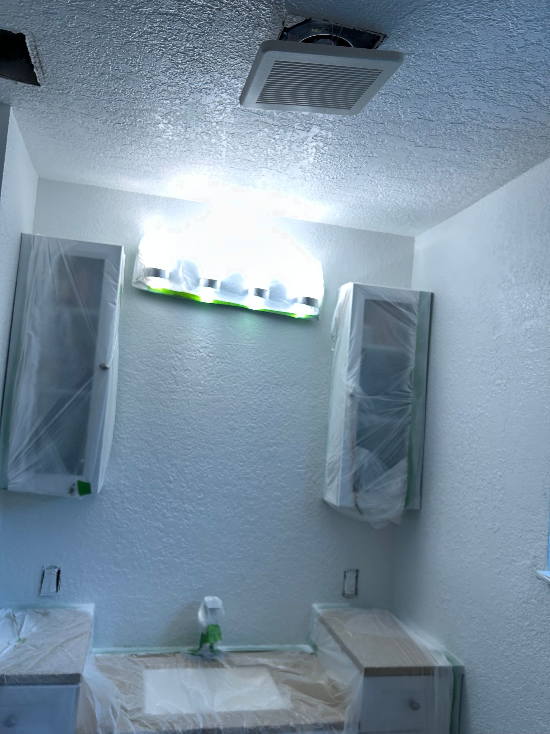 Bathroom with fresh white paint, cabinets covered in plastic, overhead lighting, and vanity.