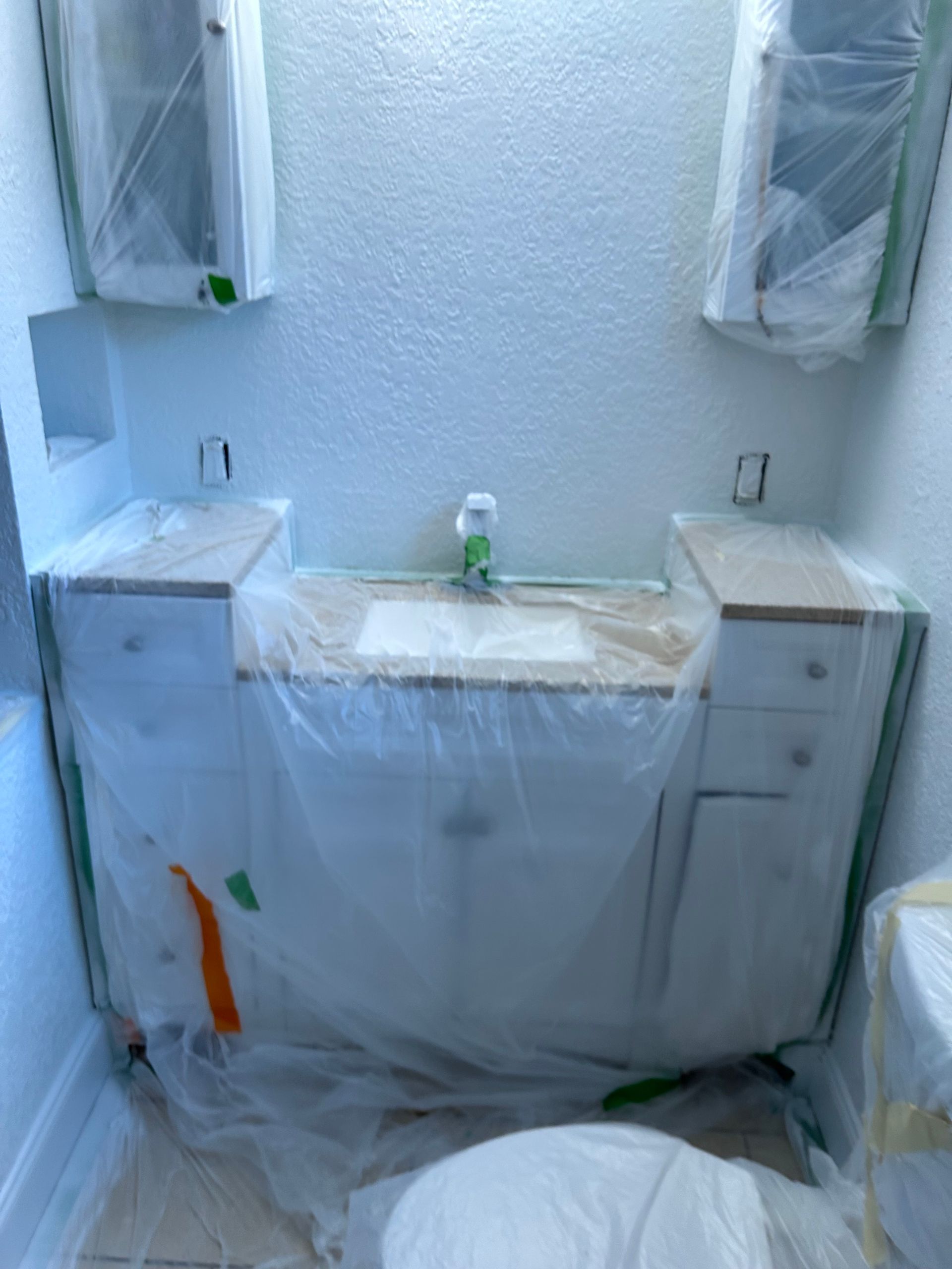 Bathroom renovation: White cabinets and countertop covered in plastic, two wall cabinets, sink, toilet.